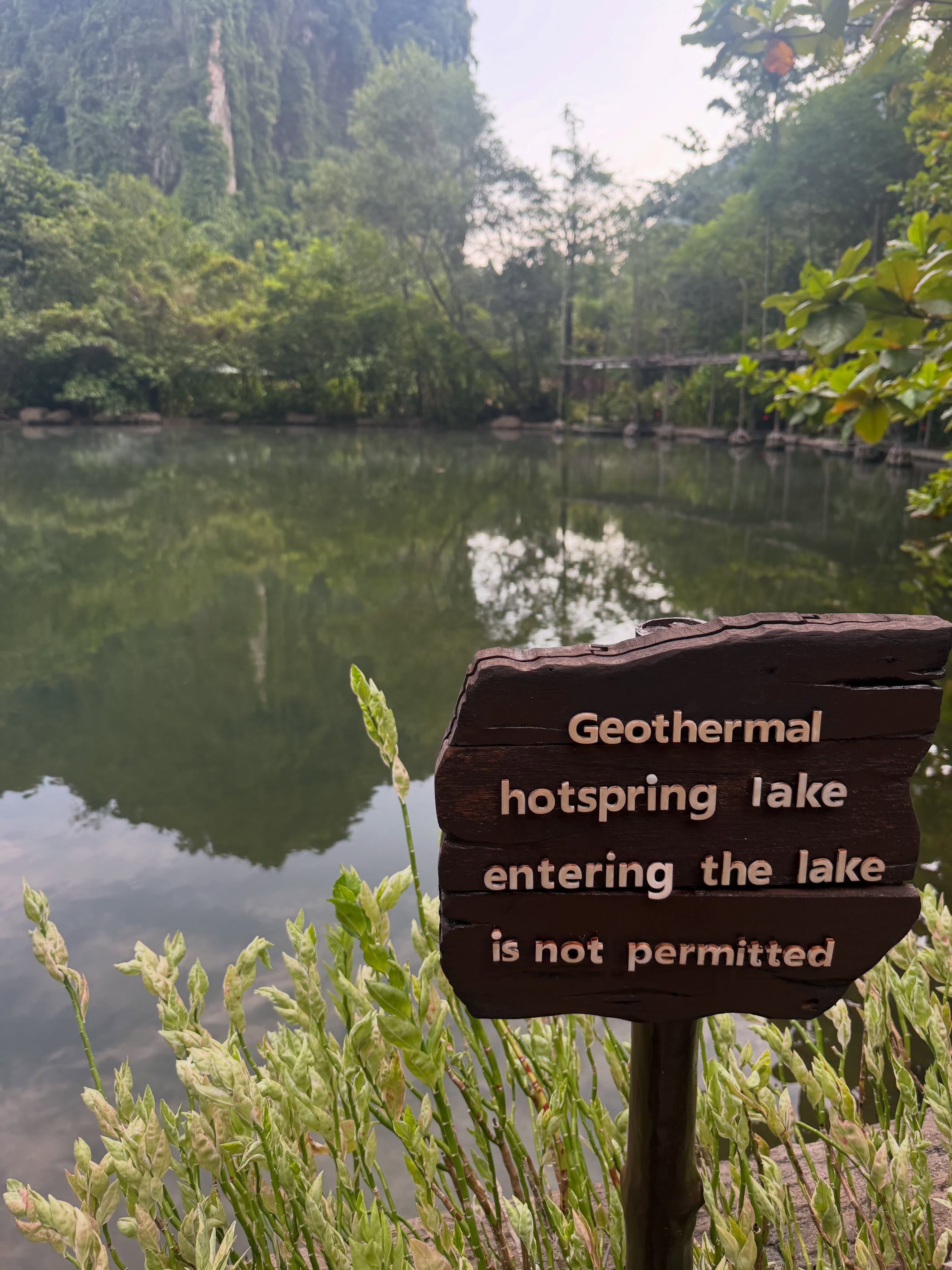 A geothermal lake beneath limestone cliffs and trees at Banjaran, with a wooden sign asking visitors not to enter the water.