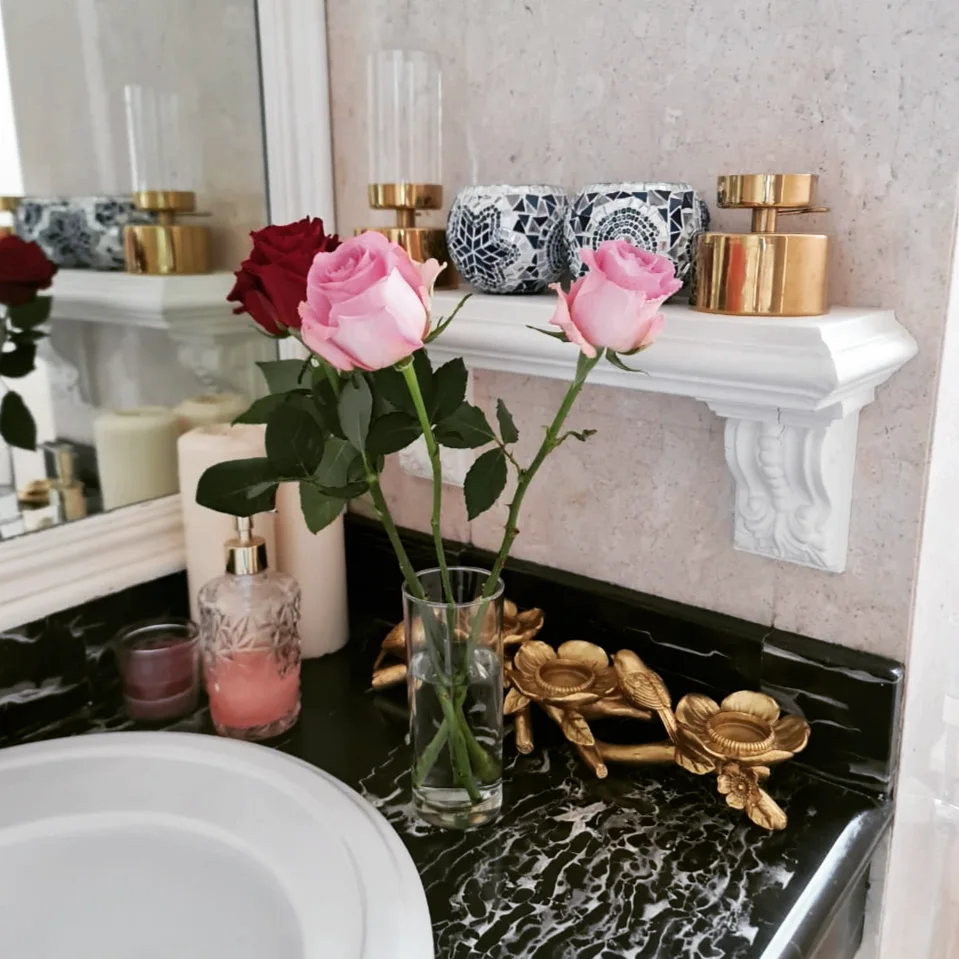 Two pink roses and a dark red rose in a glass vase on black-and-white veined marble, beside small brass flower-shaped holders.